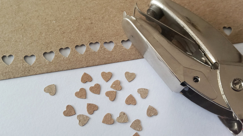 Tiny hearts cut out from brown paper lie on a tabletop next to a metal hole punch.