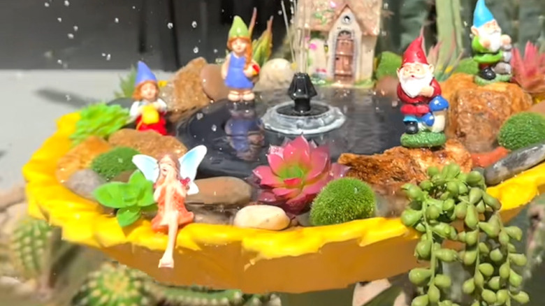 A yellow birdbath with a fountain decorated with fairy garden accessories, river stones, and fake plants.