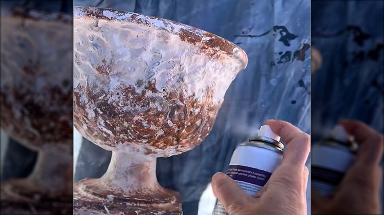 A hand holding a spray can applies paint to a thrifted urn-style ceramic planter.