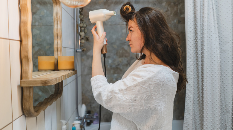 A woman blow drying her hair while using a rounded brush in a small, cramped bathroom.