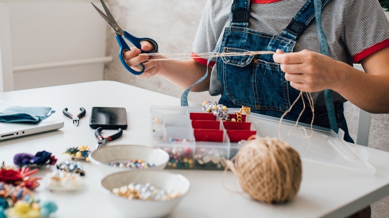A woman cuts lengths of twine with scissors while sitting at a table covered in craft supplies.