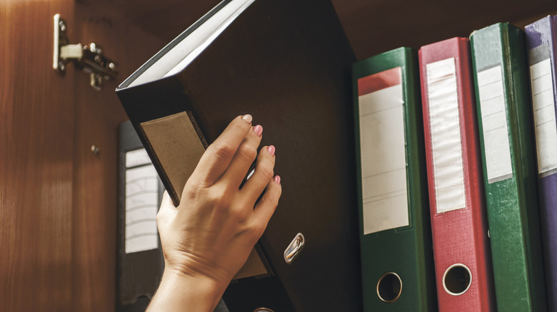 A person pulls a black binder off a cabinet shelf with numerous other binders.
