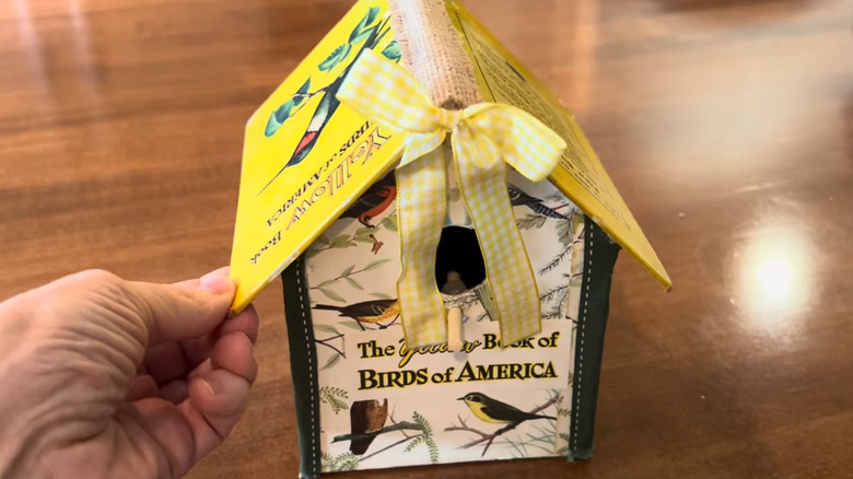 A hand moving around a birdhouse made of cardboard and a book