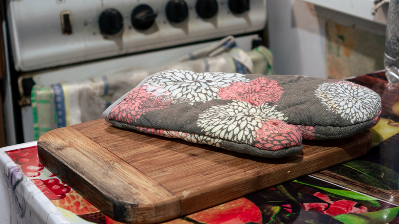 An old wooden cutting board sits on a kitchen countertop with a fabric oven mitt on it and an oven in the background.