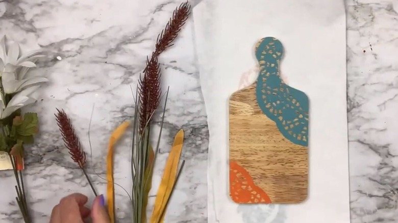 A cutting board with painted doilies and a pile of faux floral stems lying beside it on a marble countertop.