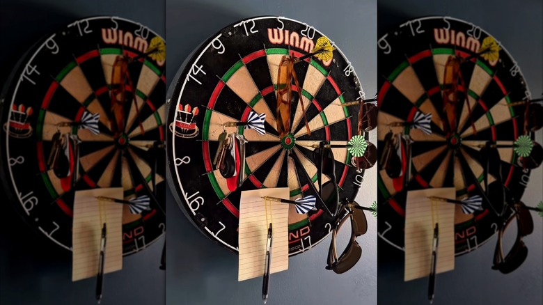 A dart board and darts being used to hang keys, sunglasses and a receipt