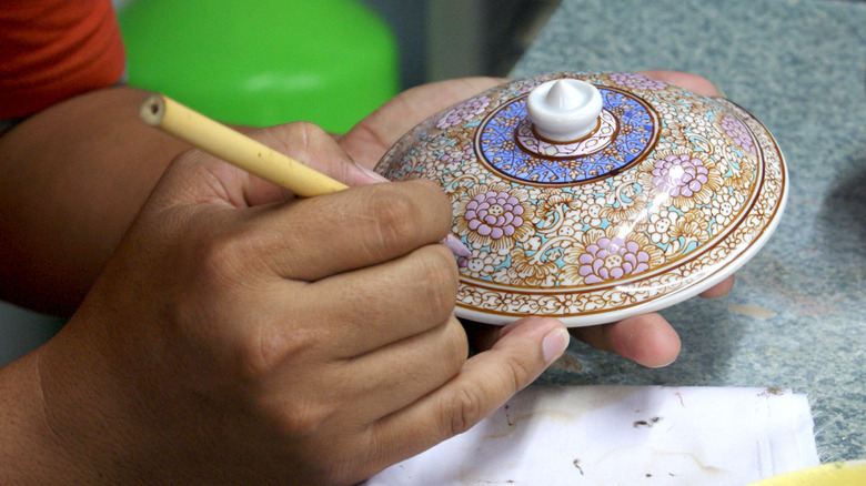 A person paints a ceramic jar lid with a bamboo paint brush.