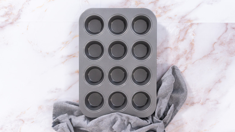 Empty muffin tin sitting on a counter with a kitchen towel.