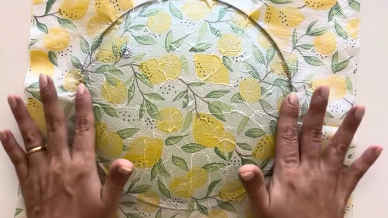 Two dark-skinned hands decoupage a glass pot lid with a patterned napkin.