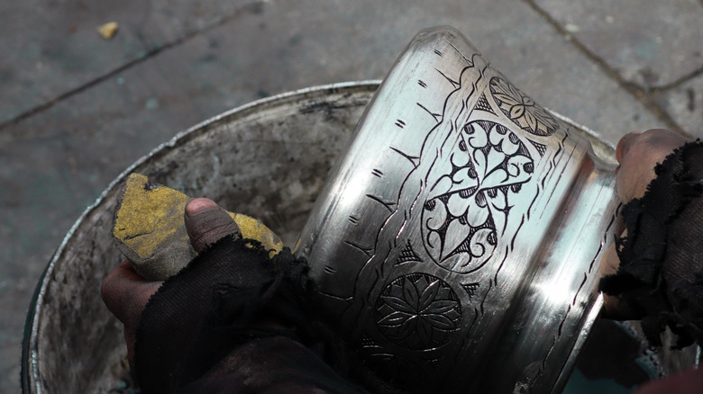 A person holds an old metal plant pot in two hands while cleaning it's surface with a sponge.