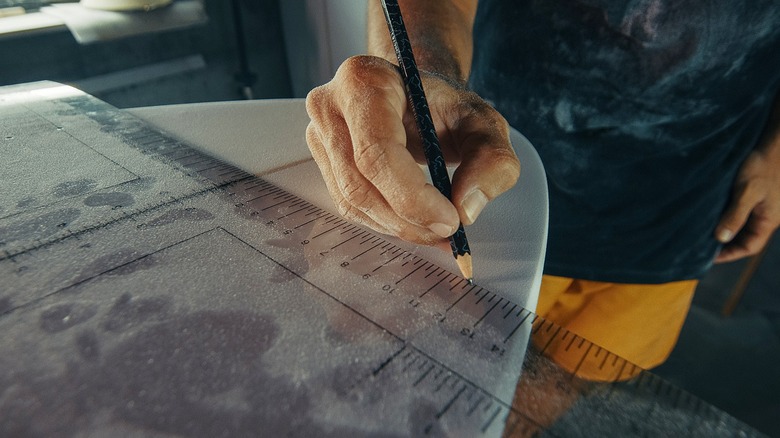 Close-up of a person's hand using a measuring tool and pencil to draw on a surfboard