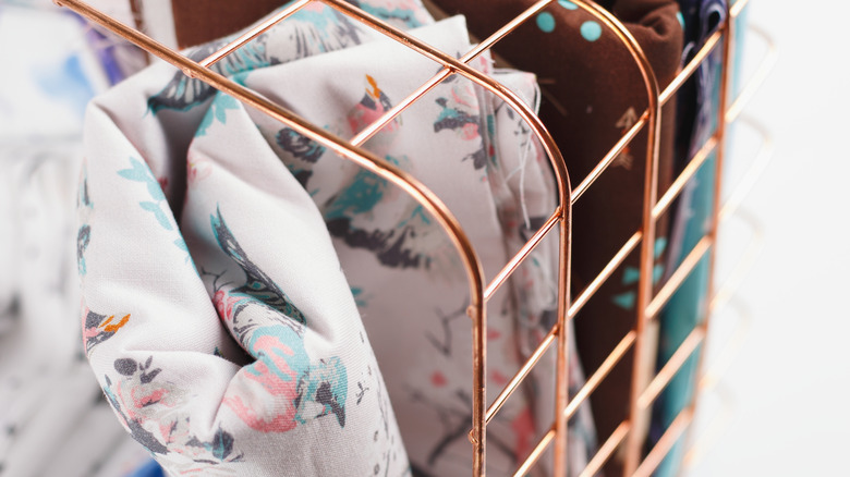 Several fabric tablecloths are contained in a bronze-colored wire basket, which stands on its side