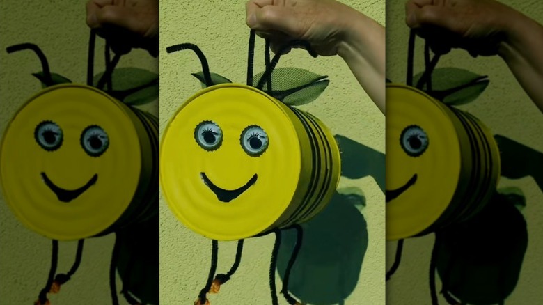 A hand holds up a hanging bee house made from a tin can painted with yellow and black stripes and adorned with a friendly face.