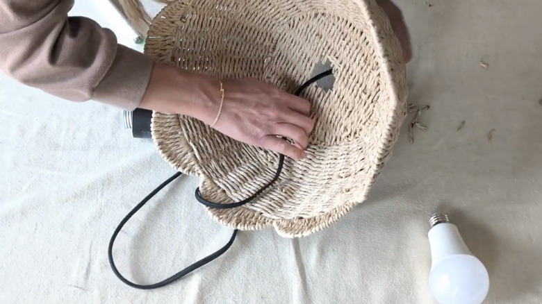A person threads a hanging light fixture through the base of a wicker basket.