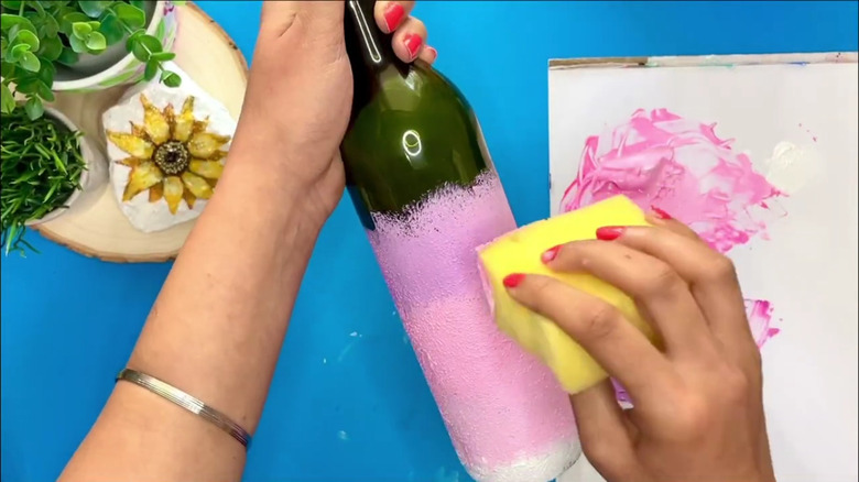 A woman's hand paints a wine bottle pink with a sponge