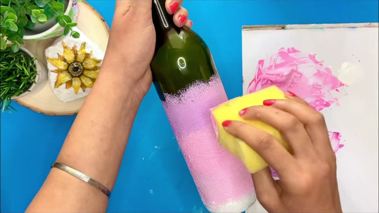 A woman's hand paints a wine bottle pink with a sponge