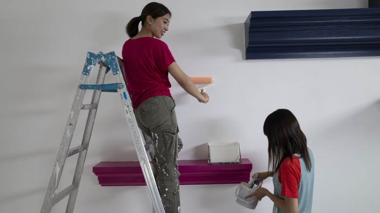 Woman on ladder painting wall around floating shelf.