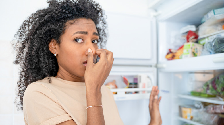 An African American woman holds her nose in front of a stinky fridge.