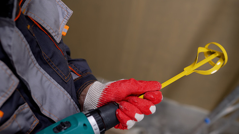 A person wearing red gloves inserts a steel paint mixer attachment into an electric drill.