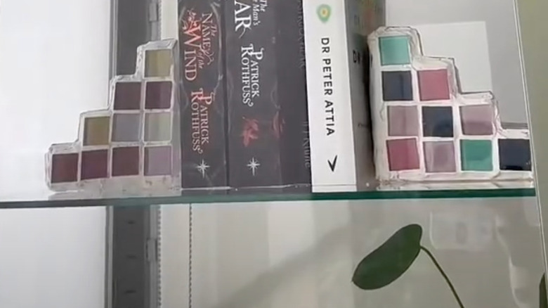 Tiled bookends with a cardboard base on a glass bookshelf supporting three books.