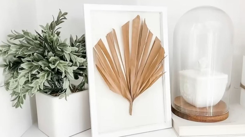 A brown leaf in a frame rests next to a plant and a figure in a glass dome.