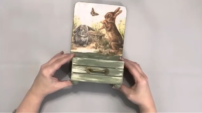 Hands pressing small wood crate onto decorated cutting board with hot glue.