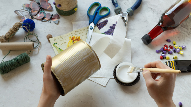 Table with craft supplies and person brushing glue onto tin can.