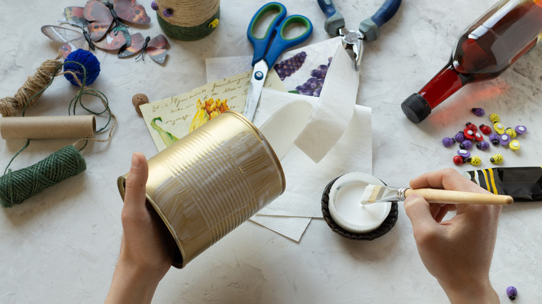 Person brushing glue on tin can at table with craft supplies
