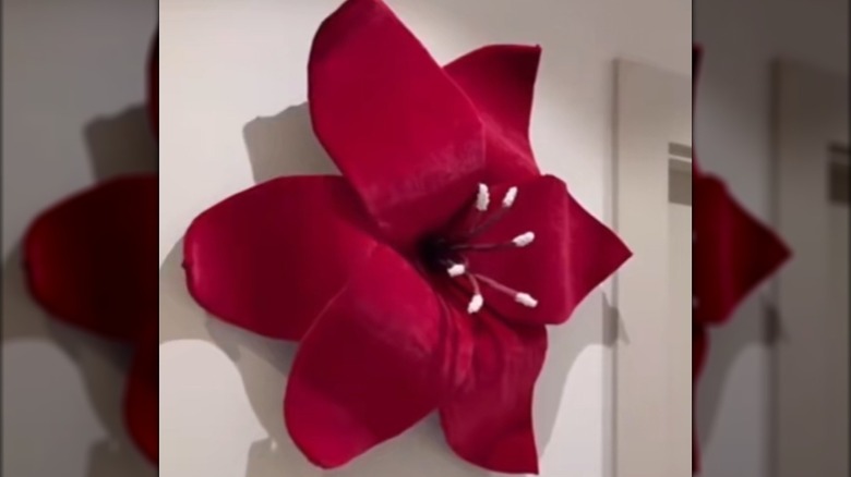 A large red flower with cloth covered cardboard petals hangs on a wall.