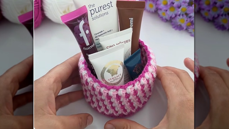 Hands holding a small crocheted basket in fuschia, light pink and white with beauty items inside it.