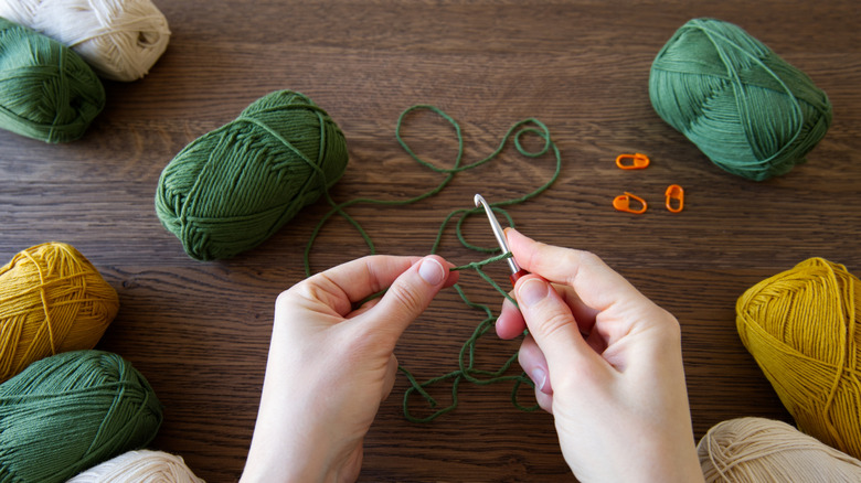 Hands crocheting with a hook and green yarn on a wooden table.