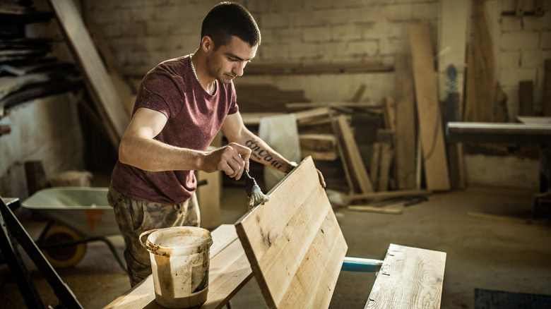 Young man stains a wood plank in a dimly lit workshop.