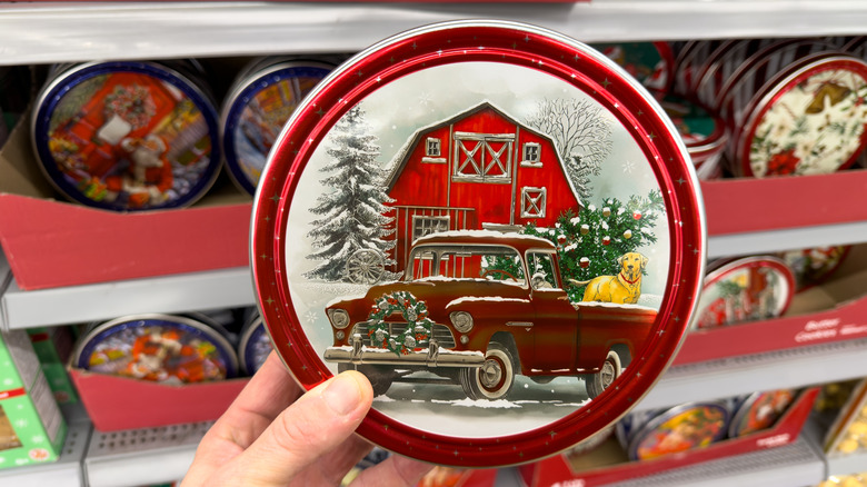 A hand holds a round cookie tin in front of other cookie tins on a store shelf.