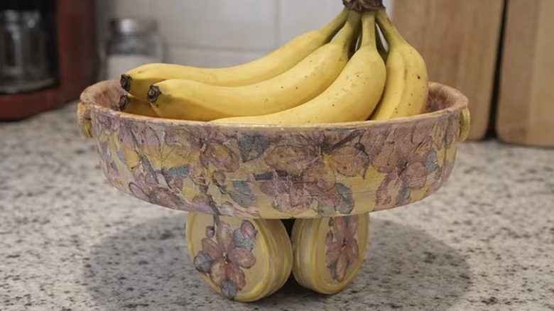 A bowl made out of a frying pan and old tins with bananas in it on a counter.
