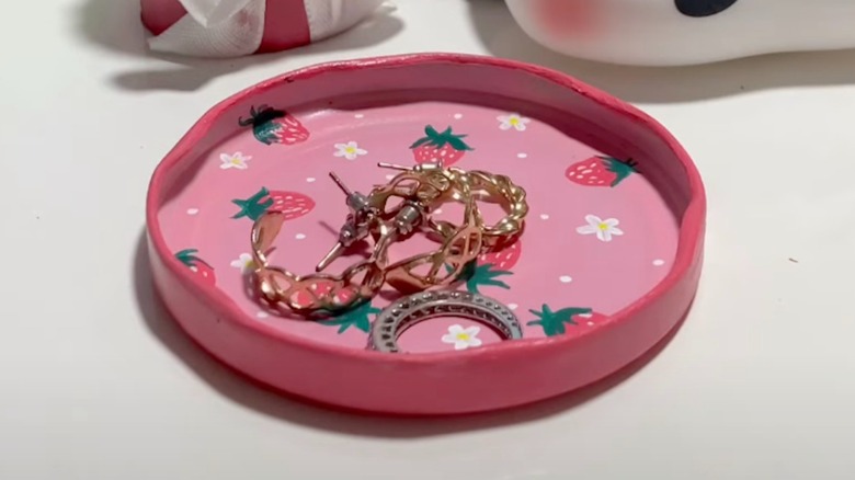 An old jar lid that has been painted pink with flowers and strawberries holding small jewelry items.