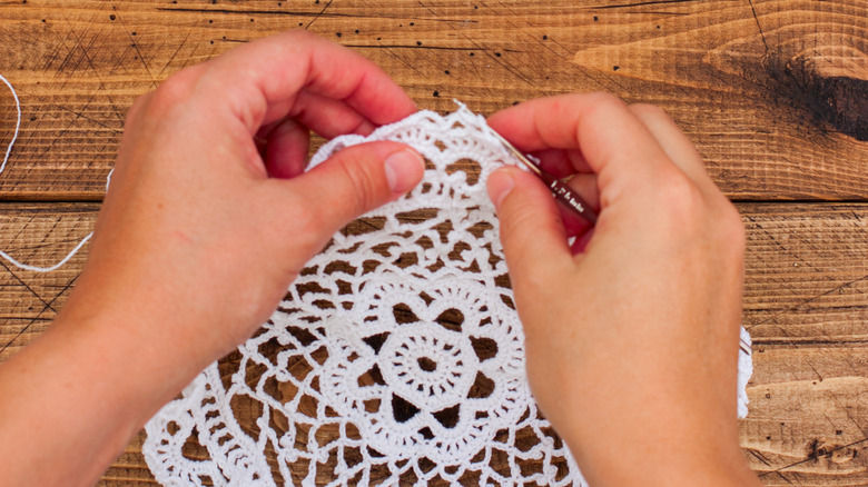 Two hands stitching a doily above a wooden tabletop.