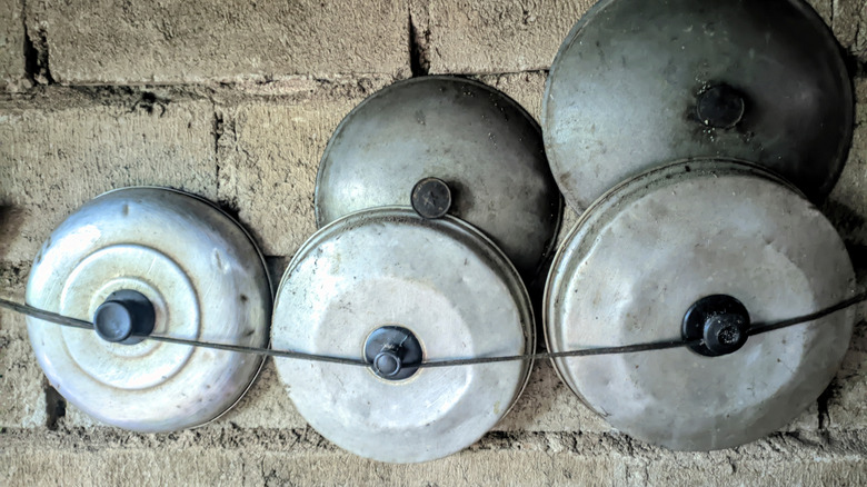 Old aluminum pot lids without their matching pots hang behind some string on a wall in a rustic kitchen.