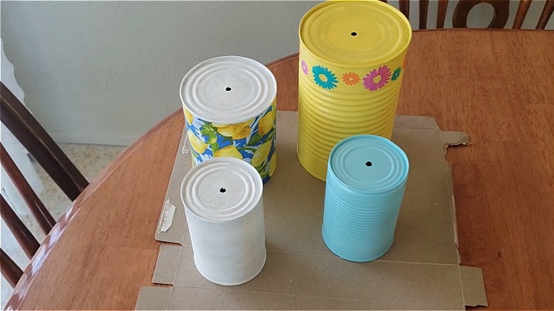 Tin cans that have been painted and decorated in bright colors are drying on a kitchen table.