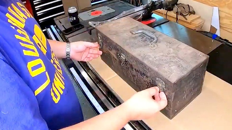 A person undoes the latches on an old toolbox sitting on a shop table