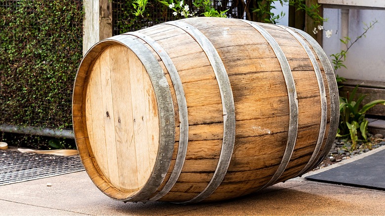 A single wine barrel sits on its side on the sidewalk with greenery seen in the background