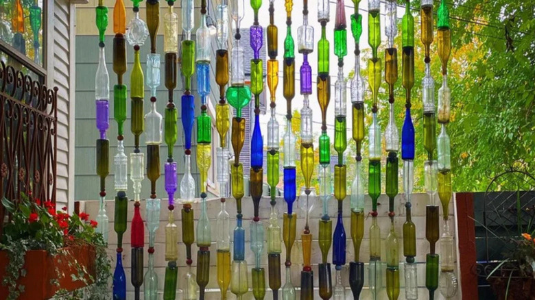 Colorful wine bottles strung vertically to create a semi-transparent outdoor privacy screen, with green, blue, yellow, and clear glass hanging in rows beside a patio and garden.
