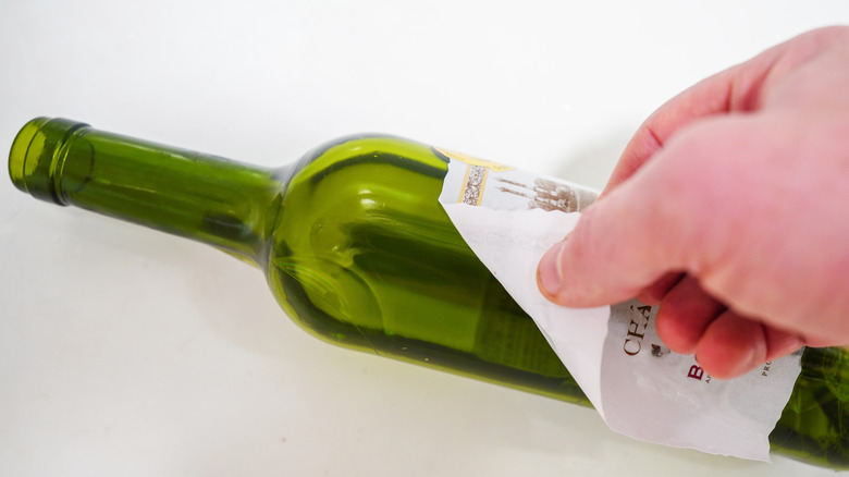 Hand peeling a paper label off a green wine bottle, showing the label being removed from the glass surface.