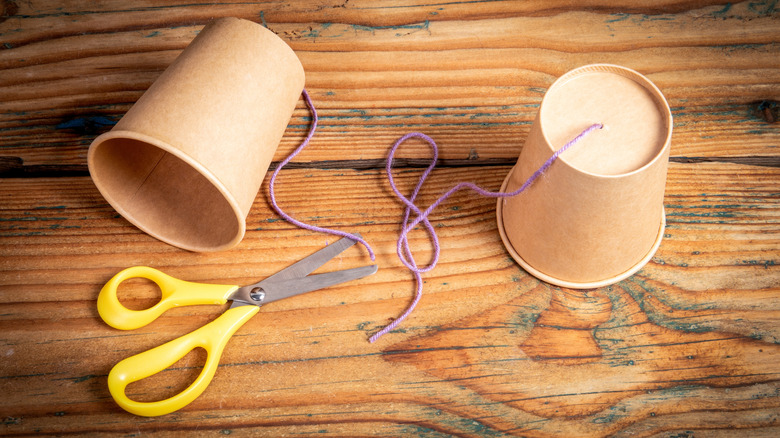 Two paper cups, string, and scissors sit on a wooden tabletop.