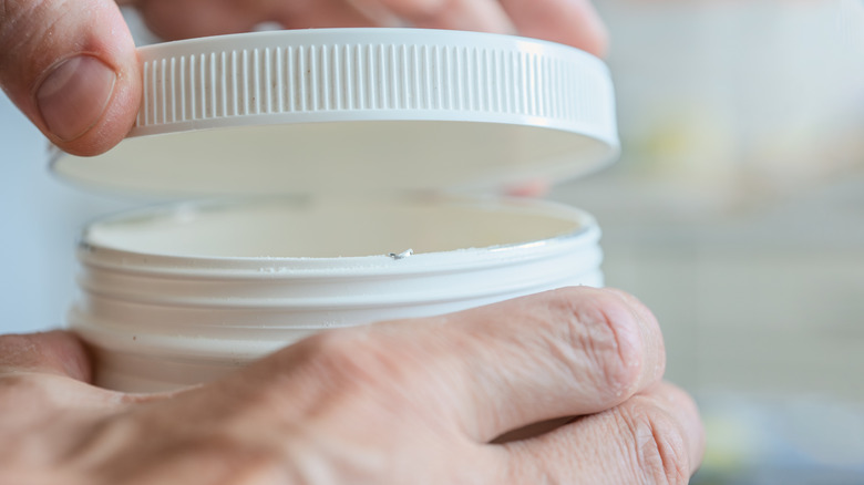 A hand removes a round plastic lid from a white plastic canister