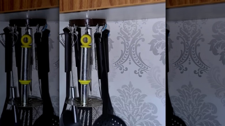 Kitchen utensils hanging from hooks attached to the plastic screw-top of a peanut butter jar affixed to the underside of an upper cabinet.