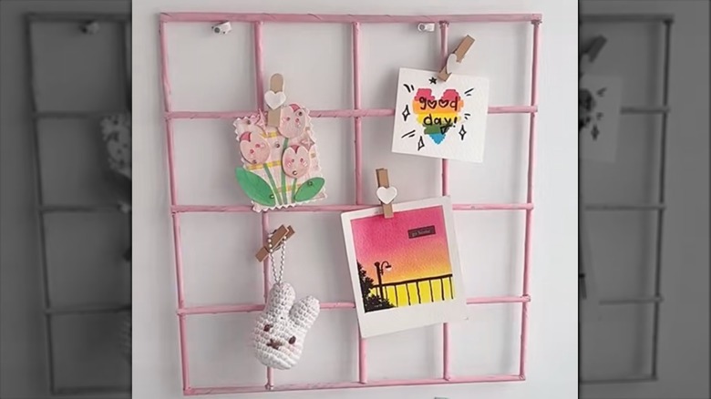 A pink wall grid with a photo, drawing, keychain, and flower craft clipped on it.