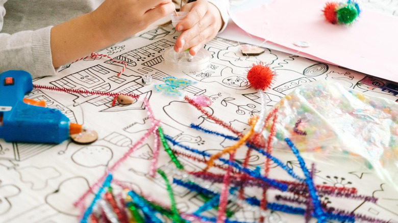 A person crafts with pipe cleaners, pom poms, and a glue gun at a table.