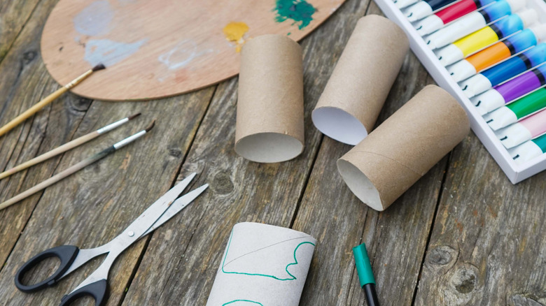 Craft tools, paint, and empty toilet paper rolls sit on a wooden tabletop.