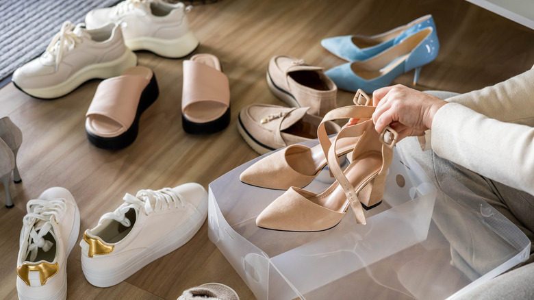 Closeup of person holding a pair of high heeled shoes, with other shoes strewn around on the floor