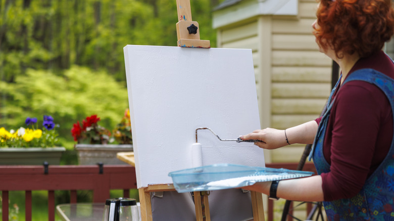 A woman outside on a deck paints a canvas on an easel white with a lightly textured paint roller.
